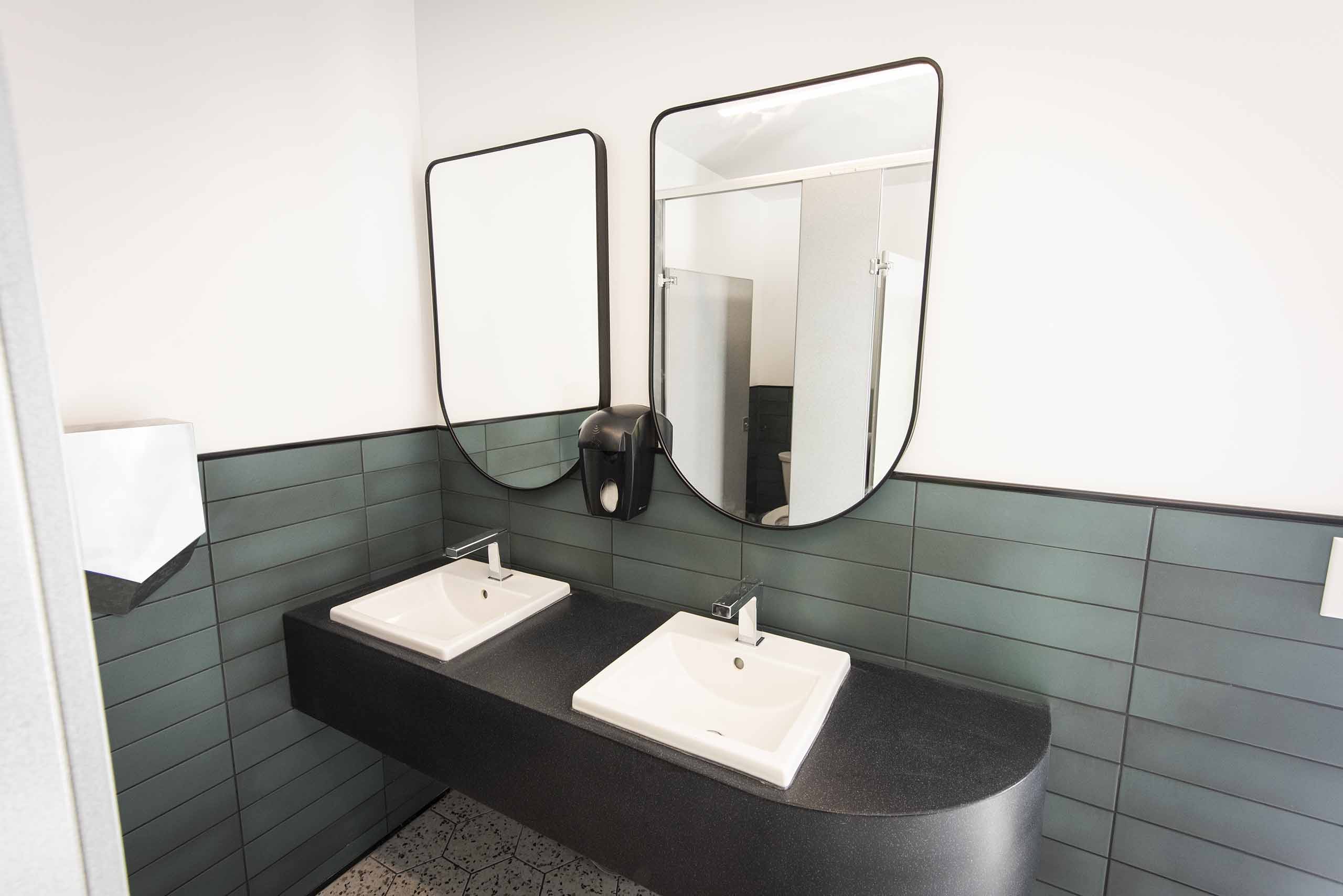 A double washroom sink with double mirrors over a white wall with green subway tile wainscotting.